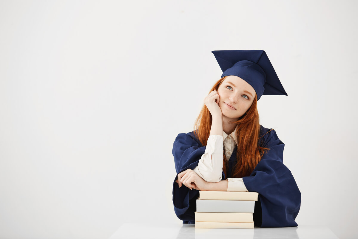 Jovem ruiva com roupa de formanda apoiada em livros pensando no medo de fazer faculdade.