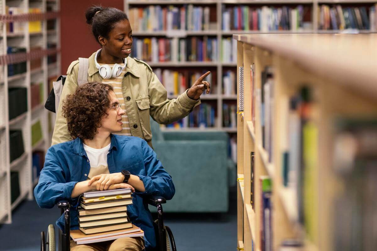 Mulher ajudando colega cadeirante em biblioteca, promovendo a acessibilidade na educação.