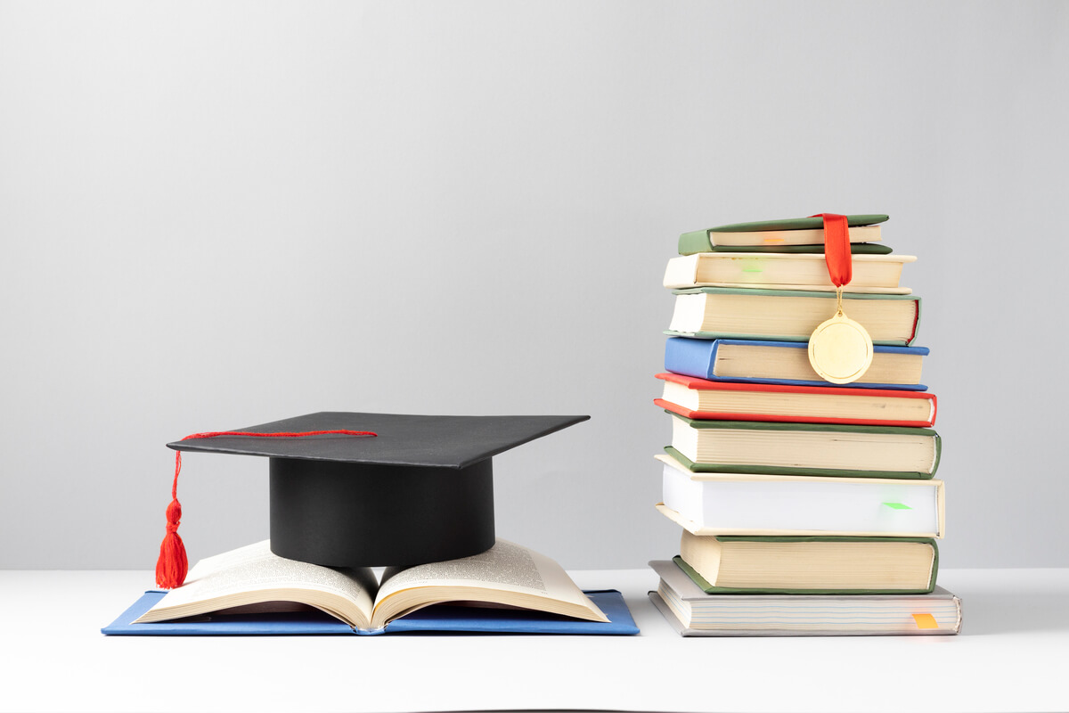 Livros empilhados, uma medalha e um chapéu de formatura de pós-graduação em outra área.