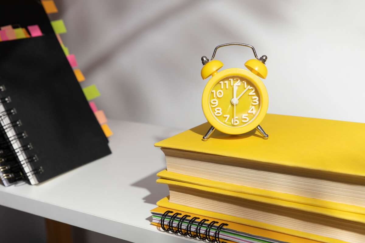Livros amarelos com um relógio em cima para contar o tempo destinado ao estudo para faculdade e concurso.
