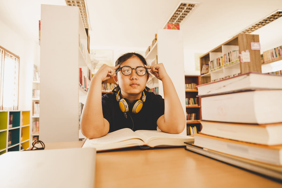 Menina asiática estudando na biblioteca com livros, pensando em como manter o foco.